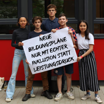 Auf dem Bild sind fünf junge Menschen zu sehen, die zusammen ein Schild halten. Auf dem Schild steht: "Neue Bildungspläne nur mit echter Inklusion statt Leistungsdruck"